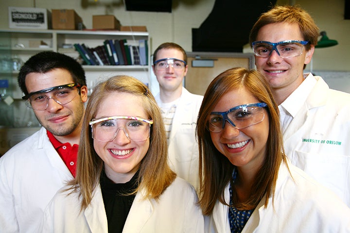 Five students wearing safety glasses and white lab coats smile for the camera. Lab shelves are in the background.